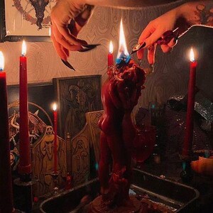 May include: A red candle sculpture of two figures embracing, with a blue flame, surrounded by lit red candles. A framed image of a goat's head is visible in the background. The scene is set in a dimly lit room.