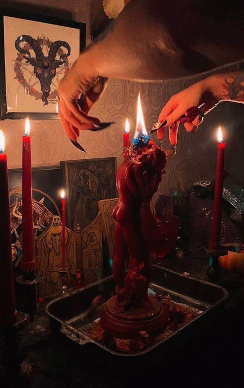 May include: A red candle sculpture of two figures embracing, with a blue flame, surrounded by lit red candles. A framed image of a goat's head is visible in the background. The scene is set in a dimly lit room.