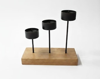 May include: A decorative candle holder featuring three black metal candle cups of varying heights, mounted on a rectangular wooden base. The candle holders are arranged in a staggered pattern, creating a visually appealing display. The wooden base has a natural wood grain.