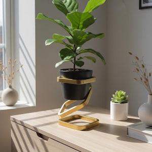 Op de afbeelding: Een levendige groene vioolbladplant in een matzwarte pot, geplaatst in een goudkleurige metalen standaard met een uniek spiraalontwerp dat de pot boven een vierkante voet verheft, wat zorgt voor een moderne en evenwichtige uitstraling.