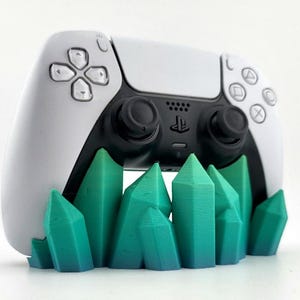 Könnte beinhalten: Ein weiß-schwarzer PlayStation 5-Controller, der auf einem teal- und grünfarbenen kristallförmigen Ständer ruht. Der Controller hat schwarze Joysticks und Tasten. Der Ständer ist ein dekorativer Artikel.