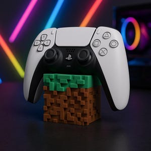 Puede incluir: Un mando blanco y negro de PlayStation 5 descansa sobre un bloque pixelado marrón y verde. El mando tiene un centro negro con laterales y botones blancos. El bloque está diseñado para parecerse a un bloque de Minecraft.