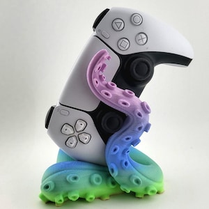 Könnte beinhalten: Ein weißer Gamecontroller wird von einem farbenfrohen, farbverlaufenden, oktopusartigen Ständer gehalten. Der Ständer wechselt von Grün zu Blau zu Lila. Der Controller hat schwarze Akzente und Tasten-Symbole.