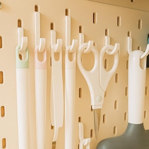 May include: A beige pegboard with various white and pastel-colored tools hanging on it. The tools include a pair of scissors, a spatula, and several other cleaning or crafting tools. The pegboard has evenly spaced holes.