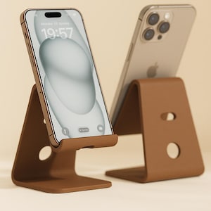 Könnte beinhalten: Zwei braune Handyhalter aus Leder, die jeweils ein Smartphone halten. Das Telefon links zeigt die Uhrzeit 19:57 und das Datum Montag, 23. Januar. Die Ständer haben ein minimalistisches Design mit einem kreisförmigen und einem tropfenförmigen Ausschnitt.
