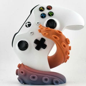 Könnte beinhalten: Ein weißer Xbox-Controller wird von einem orange-blauen, tentakelförmigen Ständer gehalten. Der Controller hat schwarze Tasten und farbige Aktionstasten. Der Ständer ist als Oktopus-Tentakel gestaltet.