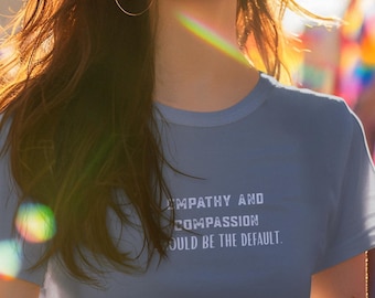 May include: A person wearing a light blue t-shirt with the text "EMPATHY AND COMPASSION SHOULD BE THE DEFAULT" in white. The background features a rainbow flag. The person is wearing gold hoop earrings.