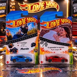 May include: Two Hot Wheels toy cars in blister packaging. The left package features a blue sports car, a motorcycle rider, and text reading "Contigo en todo el mundo". The right package features a red sports car, a couple, and "Tú ganaste la carrera por siempre".
