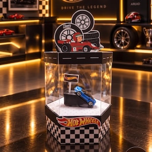 May include: A hexagonal Hot Wheels display case with clear sides and a black and white checkered base. Inside, a blue toy car sits on a black ramp with a small Hot Wheels flag. The top features a cutout of a race car driver in a red car.