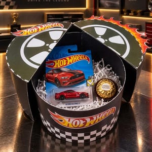 Peut inclure: Une boîte cadeau ronde noire avec un motif à damier et la marque Hot Wheels contenant une voiture miniature rouge 18 Ford Mustang GT dans son emballage bleu d'origine et un chocolat Ferrero Rocher doré niché dans des copeaux de papier blanc.