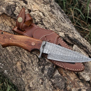 May include: A fixed-blade knife featuring a patterned Damascus steel blade and a polished wooden handle with a brass pin. The knife is accompanied by a brown leather sheath with white stitching and a brass snap button closure.
