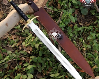 May include: A silver sword with a gold hilt and black handle rests on a wooden branch, next to a brown leather sheath. The sword has a double-edged blade. The sheath is open, revealing the sword's blade. The background is green foliage.