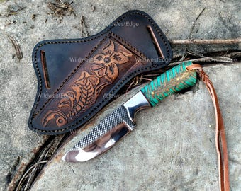 May include: A fixed-blade knife with a textured metal rasp blade, a polished edge, and a bright turquoise and brown patterned handle. It includes a brown leather sheath featuring an embossed floral design and a long leather lanyard attached.
