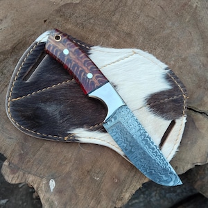 以下が含まれることがあります： 模様のある刃と赤いアクセントの木製ハンドルが付いた手作りのナイフ。ナイフは、茶色と白の模様の牛革の鞘の上に置かれています。ナイフと鞘は木製の表面に表示されています。