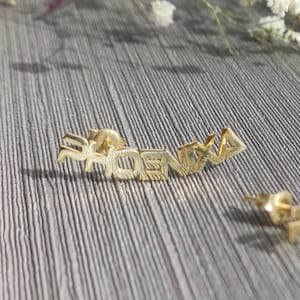 May include: A pair of gold-coloured "PHOENIX" ear climbers. The earrings are in a script font and feature a post backing. The earrings are displayed on a grey surface with white flowers in the background.