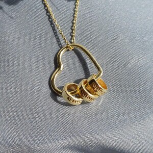 May include: A gold-colored necklace with a heart-shaped pendant. The pendant has three small rings attached, each engraved with a name. The necklace is displayed on a gray fabric surface, with white flowers in the background.