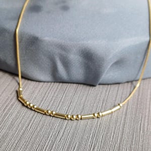 以下が含まれることがあります： 小さな円筒形と球形のビーズが連なった繊細なゴールドネックレス。 ネックレスは灰色の表面に表示され、背景には柔らかい青灰色の生地があります。 チェーンは細く、ビーズは均等に配置されています。