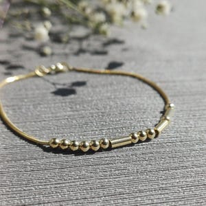 May include: A delicate gold bracelet featuring small, round beads and cylindrical accents. The bracelet is strung on a fine gold chain, creating a minimalist and elegant design. The bracelet is shown on a textured gray surface.