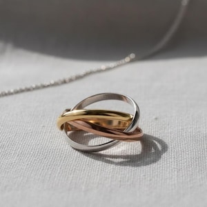 Könnte beinhalten: Nahaufnahme eines dreifarbigen Rings mit drei ineinandergreifenden Bändern in Silber, Gold und Roségold. Eine silberne Kette ist im Hintergrund leicht zu sehen, auf einer hellgrauen Oberfläche liegend.