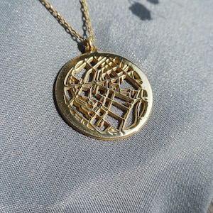 May include: A gold-colored pendant necklace with a circular, openwork design. The pendant hangs from a delicate chain. The intricate pattern within the circle creates a detailed, geometric appearance. The necklace is displayed on a gray fabric surface.
