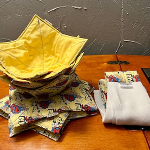 May include: A stack of yellow and patterned fabric bowl cozies with a cartoon design, alongside folded white fabric items. The bowl cozies have a star-like shape and are stacked on a wooden surface. The fabric features a yellow background with cartoon characters.