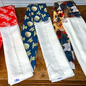 May include: Three hanging kitchen towels, each featuring a patterned fabric top section and a solid white bottom section. The patterns include a red Coca-Cola logo print, a blue Grogu character print, and a multi-colored sunflower and chicken print.