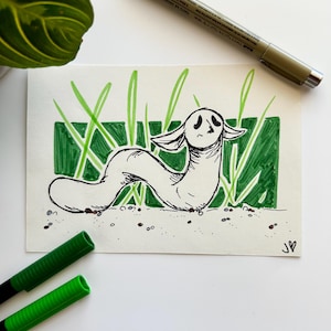 May include: An ink drawing of a white, worm-like creature with large ears and a sad face, set in green grass. The artwork is on white paper, with green markers and a pen visible. The drawing is signed with a heart.