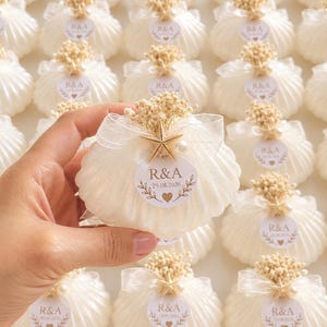 Puede incluir: Una vela blanca con forma de concha decorada con una cinta blanca transparente, una pequeña estrella de mar, una perla y flores secas. Una etiqueta circular muestra el texto R&A, la fecha 29.08.2026 y un corazón, rodeada de velas idénticas.