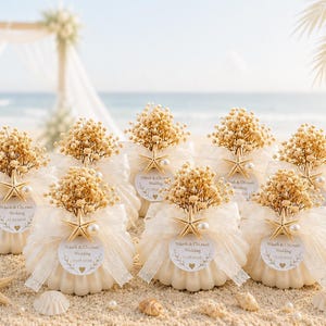 Peut inclure: Un ensemble de bougies blanches en forme de coquillage ornées de fleurs séchées gypsophile, d'un ruban en organza blanc, d'une petite étoile de mer et d'une perle. Chaque bougie porte une étiquette ronde avec le texte "Vikash & Chinu Wedding 17.07.2024" et un cœur.