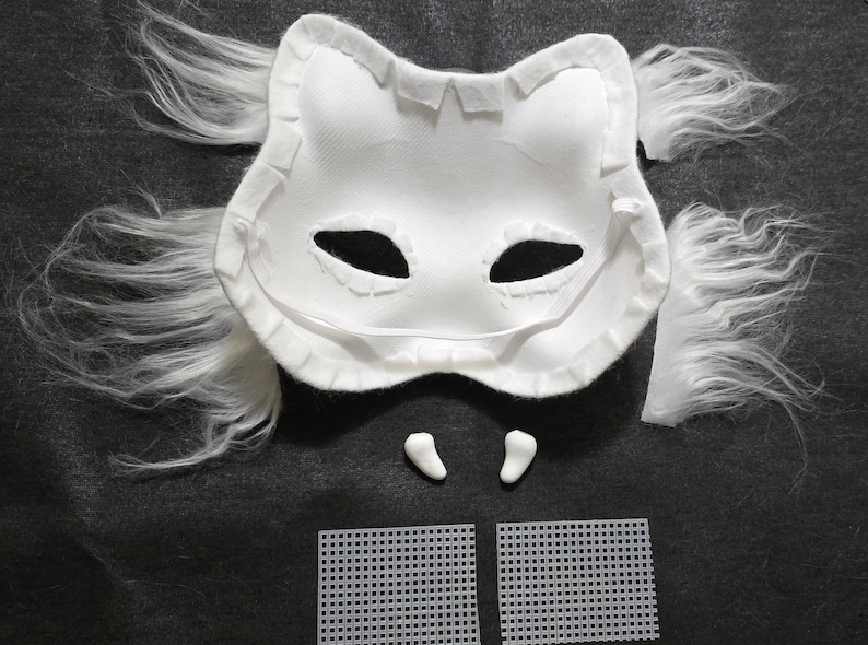 May include: A white animal-shaped mask base with attached long white faux fur tufts on the sides. The mask features two eye openings, two small white tooth-like attachments positioned below the mask, and two separate white plastic mesh squares.