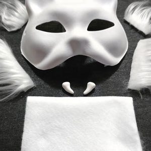 Op de afbeelding: Een wit kattenmasker met oor- en ooguitsparingen, omringd door witte imitatiebont stukken, twee kleine witte tanden, een witte vilten vierkant en een grijs elastiek. Deze knutselset is voor het maken van een kostuum of cosplay.