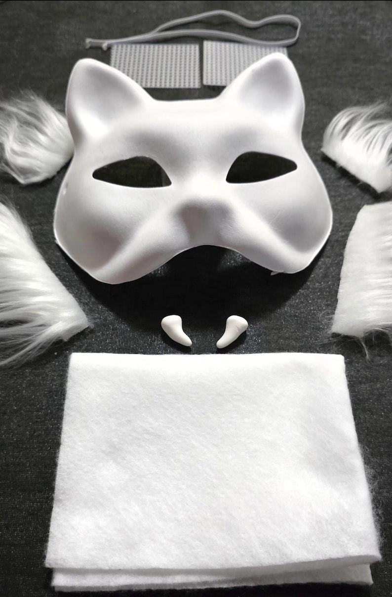 Op de afbeelding: Een wit kattenmasker met oor- en ooguitsparingen, omringd door witte imitatiebont stukken, twee kleine witte tanden, een witte vilten vierkant en een grijs elastiek. Deze knutselset is voor het maken van een kostuum of cosplay.
