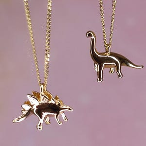 Könnte beinhalten: Zwei goldfarbene Halsketten mit Dinosaurier-Anhängern. Ein Anhänger ist als Stegosaurus mit markanten Rückenplatten geformt, während der andere einen langhalsigen Brachiosaurus darstellt. Beide Anhänger hängen an zierlichen goldfarbenen Ketten.