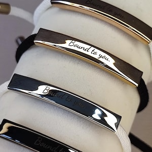 Puede incluir: Cuatro pulseras con barras metálicas rectangulares con la inscripción "Bound to you". Las barras son doradas, plateadas y negras, unidas a cordones blancos y negros. Las pulseras se exhiben sobre una superficie cilíndrica blanca.