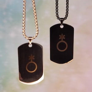 Op de afbeelding: Twee kettingen met dog tags met genderqueer symbolen. Eén tag is zwart met een gouden ketting, de andere is goudkleurig met een gouden ketting. Het genderqueer symbool is een combinatie van de mannelijke en vrouwelijke symbolen.