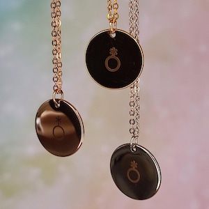 Op de afbeelding: Drie kettinghangers met ronde bedels en kettingen. De bedels zijn goud, zilver en roségoud, elk met een symbool. De kettingen zijn delicaat en hangen tegen een zachte, kleurrijke achtergrond.