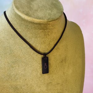 Könnte beinhalten: Eine schwarze Anhängerhalskette mit einem rechteckigen Anhänger und einer dunkelbraunen Kordel. Der Anhänger zeigt ein rosa Symbol. Die Halskette wird auf einer Schaufensterpuppe mit grünem Stoff präsentiert.