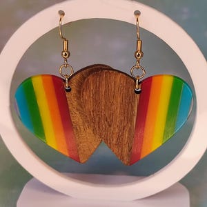 Op de afbeelding: Hartvormige oorbellen met een regenboogontwerp en goudkleurige haken. De oorbellen worden gepresenteerd op een witte standaard met een cirkelvormig ontwerp. De oorbellen hebben een houten achterkant.