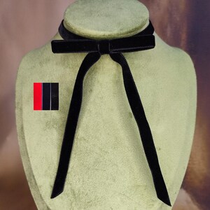 Könnte beinhalten: Ein schwarzes Samt-Choker-Halsband mit Schleife und langen Bändern, präsentiert auf einer grünen Mannequin. Ein kleiner Anstecker mit roten und schwarzen Streifen ist ebenfalls zu sehen.