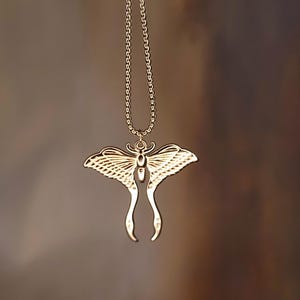 May include: A gold-coloured pendant necklace with a moth design. The moth pendant has detailed wings and a delicate chain. The necklace is shown against a blurred, neutral background.