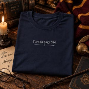 Peut inclure: Un t-shirt bleu marine foncé avec le texte blanc "Turn to page 394." centré sur la poitrine. Le texte est accompagné d'une ligne horizontale avec un petit point au milieu, positionnée directement sous la phrase imprimée.