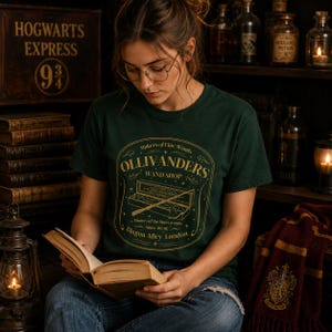 May include: A dark green short-sleeve t-shirt featuring a gold graphic print of the Ollivanders Wand Shop logo. The design includes the text Makers of Fine Wands, Ollivanders Wand Shop, Diagon Alley London, and an illustration of a wand in a box.