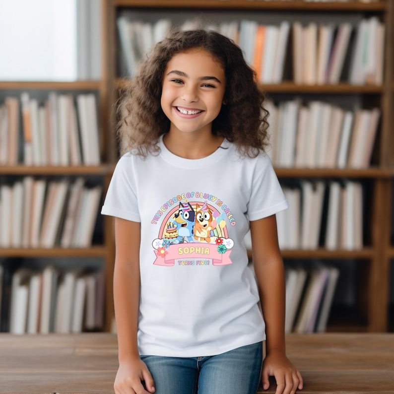 A young girl with curly hair is smiling and wearing a white t-shirt with a graphic design featuring two cartoon dogs.