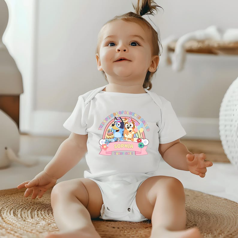 A smiling baby girl wearing a white onesie with a cartoon character design.
