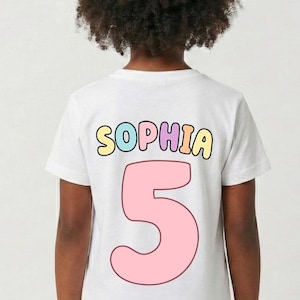 A child with curly hair wearing a white t-shirt with the name "Sophia" and the number "5" printed on it.