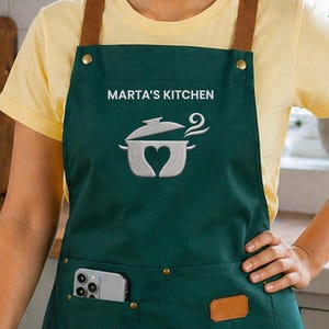 Op de afbeelding: Een donkergroene schort met bruine leren bandjes en zakken. De schort heeft de tekst "MARTA'S KITCHEN" boven een potafbeelding met een hartvorm. Een smartphone zit in de voorzak.