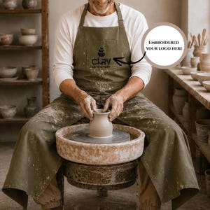 Op de afbeelding: Een persoon die een groene schort draagt met het woord "Clay" en een logo, werkt op een pottenbakkerswiel. De schort is over een wit shirt met lange mouwen. De persoon maakt een keramische vaas. Schappen met aardewerk staan op de achtergrond.