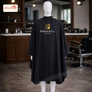 Puede incluir: Una capa de salón negra con el texto dorado "Salford & Co. HAIR SALON". La capa se muestra en un maniquí en un salón. Está diseñada para proteger la ropa durante los servicios de peluquería.