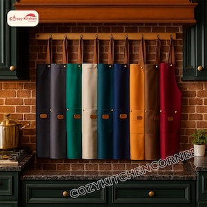 May include: A range of aprons in colours such as black, grey, green, beige, teal, navy, orange, brown, and burgundy, suspended from a wooden rack. Each apron features a leather strap and a pocket. The aprons are set against a brick wall.