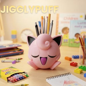 Puede incluir: Un organizador de escritorio rosa con forma de Jigglypuff que contiene bolígrafos y lápices. El organizador tiene orejas negras y una boca morada. La palabra "JIGGLYPUFF" se muestra arriba. También se ven crayones, un bloc de notas y bloques de construcción.